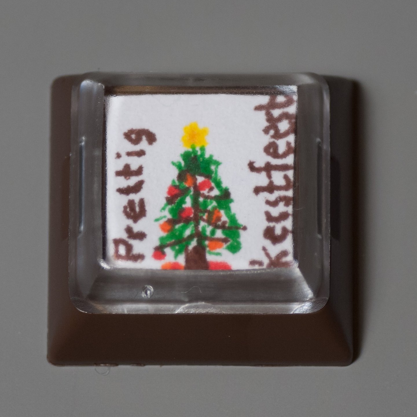 This is a relegendable DSA keycap. All craftsmanship is of the highest qualitiy. The base is shot in brown ABS, the top is molded from transparent polycarbonate. Embedded in between is a masterfully decorated piece of paper by Secret Santa.<br />On the item is an image of a Christmas tree in brown and green. The tree is decorated with red and orange balls. It is menacing with a yellow, star-formed spike. The item is inscribed with the words &quot;prettig kerstfeest&quot; in brown ink.<br />The artwork relates to the celebrating of Christmas in the Netherlands in late December of 2013.